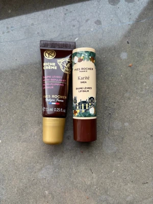 Yves Rocher läppbalsam duo - Två läppbalsam från Yves Rocher: ett i tub med guldlock, mörkbrun och guldig design, och ett i stift med beige och brun hylsa samt illustrationer. Ena varianten är Riche Crème Nutri-Plumping och den andra Karité Shea. Perfekt för återfuktade läppar.