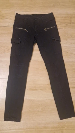 Svarta cargobyxor med dragkedjor - Säljer ett par svarta cargobyxor med smal passform och snygga dragkedjor framtill. Byxorna har klassiska cargofickor på sidorna och är tillverkade i ett mjukt, stretchigt material. Perfekta för en avslappnad och trendig stil.