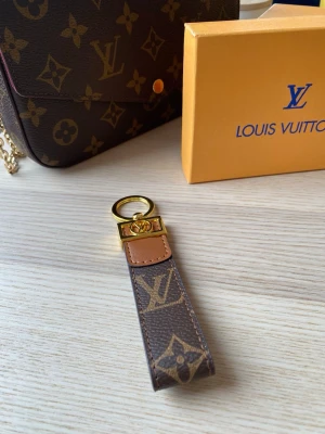 Vuitton nyckelring monogram - Snygg nyckelring från Louis Vuitton i klassiskt brunt monogrammönster med guldfärgade metalldetaljer och LV-logga. Tillverkad i canvas och läder med exklusiv känsla. Perfekt accessoar för att piffa upp väskan eller nyckelknippan.
