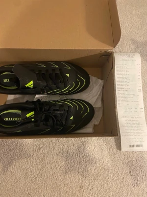 Adidas svarta fotbollsskor med neon - Säljer ett par svarta Adidas fotbollsskor med neonfärgade detaljer och snörning. Skorna har en låg profil och mönstrad sula med fasta dobbar för bra grepp på planen. Tillverkade i syntetmaterial och designade för snabbhet och kontroll.