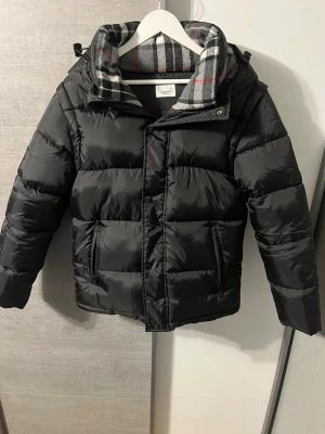 Svart pufferjacka med huva/väst - Snygg svart pufferjacka med stor huva och rutigt foder i grått, vitt och rött. Jackan har dragkedja och tryckknappar framtill samt två sidofickor. Perfekt för kalla vinterdagar och ger en riktigt chill look.