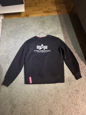 Svart sweatshirt Alpha Industries - Säljer en svart sweatshirt från Alpha Industries med deras ikoniska logga och text i vitt på bröstet. Tröjan har rund hals, långa ärmar och ribbade muddar. Klassisk och stilren design med en röd detalj vid nederkanten.