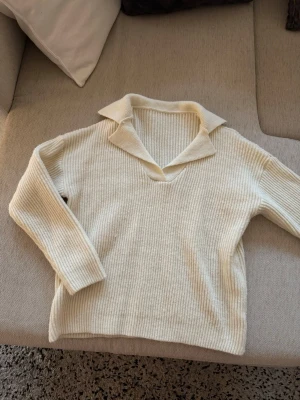 Krämvit stickad tröja med krage - Säljer en krämvit stickad tröja med bred krage och v-ringning. Tröjan har ribbad struktur och långa ärmar, perfekt för lager på lager. Materialet ser mjukt och mysigt ut, och färgen är lätt att matcha till olika outfits.