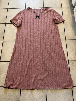 Rosa ribbat klänning med rosett storlek 1XL - Mysigt klänning i mjuk, ribbad rosa trikå med korta ärmar. Dekorativ svart rosett vid halsringningen och svarta kanter vid ärmslut och hals. Lätt utställd passform och skön stretch.