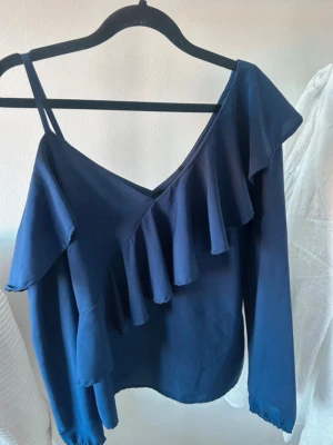 Mörkblå blus med volanger - Trendig mörkblå offshoulder blus med långa ärmar och bred volangdetalj framtill. Blusen har en axelbandsdetalj på ena sidan och en djup v-ringning. Perfekt för dig som gillar en unik och stilren look.