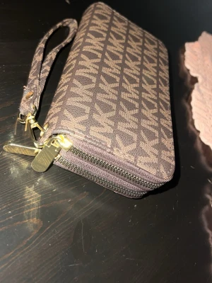 Brun Michael kros handväska  - Snygg brun plånbok från Michael Kors med klassiskt MK-logomönster över hela ytan. Plånboken har guldfärgade dragkedjor och en avtagbar handledsrem. Tillverkad i tåligt konstläder och har en rektangulär form som rymmer både kort och kontanter.