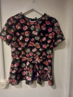 Blommig topp från Divided - Snygg blommig topp från Divided med svart botten och färgglada blommor i rosa, rött och vitt. Modellen har korta ärmar och lätt utställd nederkant som ger en feminin siluett. Perfekt för dig som gillar romantiska mönster och vill sticka ut.
