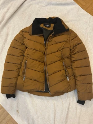 Brun pufferjacka från Sweet - Säljer en brun pufferjacka från Sweet med svart krage och foder. Jackan har två stora fickor med dragkedja framtill och en rejäl dragkedja i mitten. Puffig modell som håller dig varm under kalla dagar. Perfekt för dig som vill ha en stilren och trendig vinterjacka.