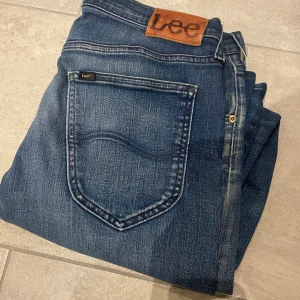 Blå Lee jeans med raka ben - Säljer ett par klassiska blå jeans från Lee med raka ben och snyggt slitna detaljer. Jeansen har fem fickor, Lee-logga på bakfickan och läderpatch i midjan. Materialet är robust denim som ger en cool vintagekänsla.