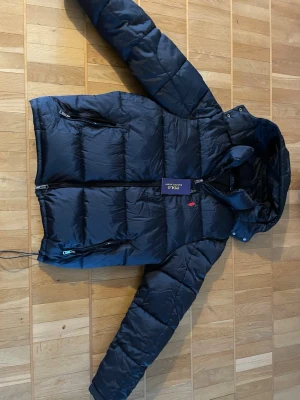 Svart dunjacka från Polo Ralph Lauren - Helt ny utan prislapp, Svart dunjacka från Polo Ralph Lauren med huva och dragkedja framtill. Med avtagbar luva. Jackan har två sidofickor med dragkedja och en bröstficka. Klassisk pufferstil med diskret röd logga på bröstet och quiltad design. Perfekt för kalla dagar.
