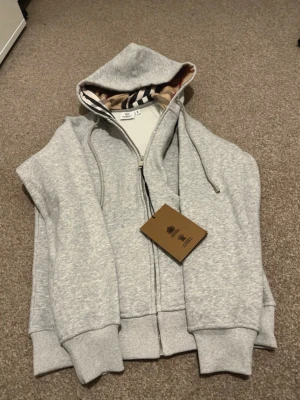 Grå zip hoodie från Burberry - Snygg grå zip hoodie från Burberry med klassiskt rutigt foder i huvan och dragsko. Tillverkad i mjuk bomullsblandning, har ribbade muddar och fickor framtill. Perfekt för en chill och stilren look.
