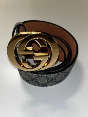  GG bälte med guldspänne - Snyggt GG bälte med klassiskt GG-mönster i grått och svart. Bältet har ett stort, glänsande guldspänne med GG-logga och insida i brunt skinn. Perfekt accessoar för att lyfta vilken outfit som helst.