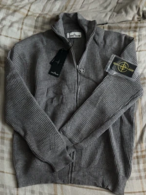 Grå stickad ziptröja från Stone Island - Säljer en grå stickad ziptröja från Stone Island med ribbad struktur och hög krage. Tröjan har dragkedja med ikonisk kompassdetalj och avtagbart märke till ärmen. Perfekt för dig som gillar streetwear och snygga detaljer.