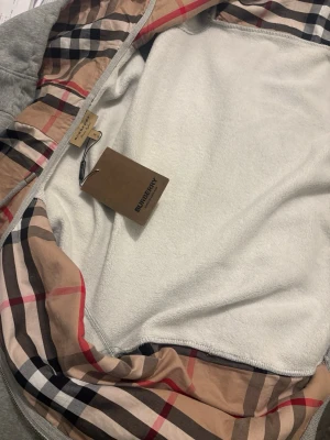 Grå zip hoodie från Burberry - Säljer en grå zip hoodie från Burberry med klassiskt rutigt foder i huvan och diskret logga på bröstet. bästa kvaliten du kan hitta i plick.  Passar många outfits och kan användas på både vintern och sommarn
