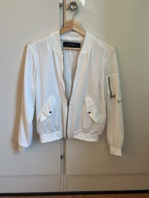 Vit bomberjacka från Zara Basic - Snygg vit bomberjacka från Zara Basic med dragkedja framtill, två fickor med tryckknappar och en ärmficka med dragkedja. Jackan har ribbade muddar vid ärmslut och midja samt en klassisk bomberkrage. Materialet är tunt och ger en stilren look. Dragkedjan är lite dåligt men den fungera fortfarande.