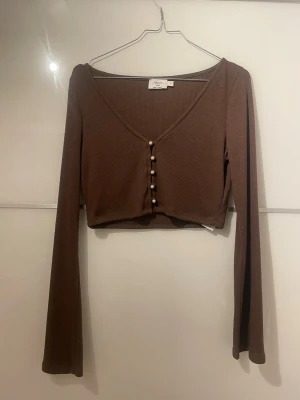 Brun croppad topp från NA-KD - Snygg brun croppad topp från Hanna Scönbergs kollektion med nakd. Långa ärmar och v-ringning. Toppen har ribbad struktur och dekorativa pärlknappar framtill. Perfekt att styla med höga jeans eller kjol.