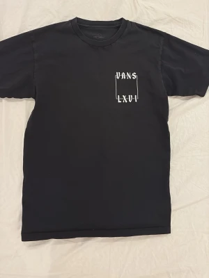 Svart Vans t-shirt med ormtryck - Säljer en svart t-shirt från Vans med coolt vitt tryck fram och stort ormtryck med texten 'Vans The Original LXVI 66' på ryggen. Klassisk passform och rund hals. Perfekt för dig som gillar streetwear och snygga detaljer.