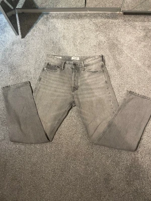 Grå raka jeans från Jack & Jones - Säljer ett par grå jeans från Jack & Jones med rak passform och klassisk femficksdesign. Jeansen har en ljusgrå tvätt och är tillverkade i bomull. Perfekta för en avslappnad och trendig look.