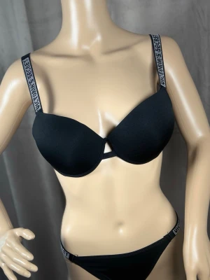 Svart push-up bh från Victoria's Secret 70D - Snygg svart push-up bh från Victoria's Secret med vadderade kupor och dekorativa axelband med logotyp i vitt. Bh:n har en liten cut-out detalj framtill och justerbara axelband för bästa passform. Perfekt för dig som vill ha både komfort och stil.