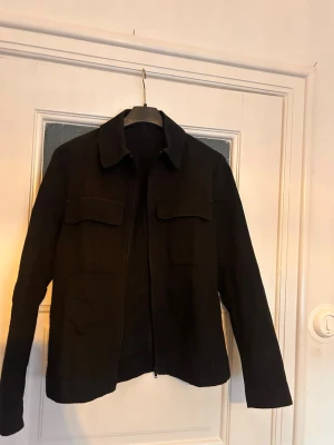 Svart overshirt från Whyred - Stilren svart overshirt från Whyred med klassisk krage, två bröstfickor och dragkedja framtill. Tillverkad i ett mjukt tyg som ger en clean look. Perfekt att bära över en t-shirt eller hoodie för en avslappnad men snygg stil. Storleken sitter som S.