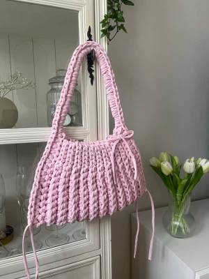 Handgjord rosa virkad handväska - Supersöt handväska i ljusrosa, virkad med tjockt garn för en chunky look. Väskan har en bågformad siluett, bred axelrem och dekorativa knytband på sidorna. Perfekt för dig som gillar unika accessoarer och vill sticka ut med en färgglad detalj.