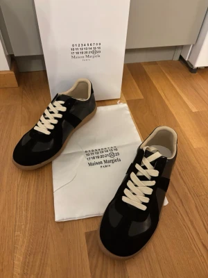 Maison Margiela svarta sneakers - Svarta sneakers från Maison Margiela med vita snören och beige innersula. Skorna har en klassisk låg silhuett och gummisula. Materialet är en mix av läder och mocka som ger en lyxig känsla. Perfekta för dig som gillar stilrena och tidlösa sneakers.