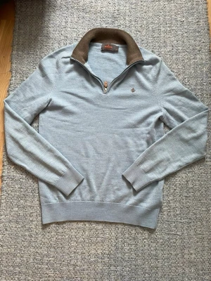 Ljusblå stickad tröja från Morris - Ljusblå stickad tröja från Morris med halv dragkedja och brun kontrastkrage. Tröjan har ett broderat brunt emblem på bröstet och är gjord i 100% merino ull den passar dig som är runt 165 cm lång