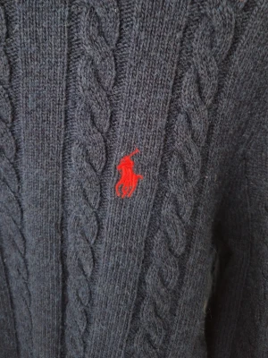 Mörkblå kabelstickad tröja Polo Ralph Lauren - Kabelstickad tröja i mörkblått från Polo Ralph Lauren med klassisk röd broderad logga på bröstet. Tröjan har grovstickat mönster och ribbade muddar. Perfekt för dig som gillar tidlös och sportig stil.