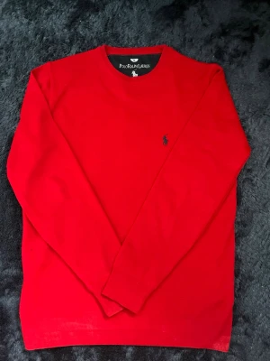 Röd stickad tröja Polo Ralph Lauren - Klassisk röd stickad tröja från Polo Ralph Lauren med rund halsringning och diskret svart logga broderad på bröstet. Tröjan har långa ärmar och ribbade muddar vid ärmslut och nederkant. Perfekt för en stilren och enkel look.