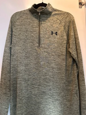 Grön träningströja från Under Armour - Snygg grönmelerad träningströja från Under Armour med halv dragkedja och hög krage. Tillverkad i ett tunnt mjukt och stretchigt funktionsmaterial som andas. Perfekt för löpning eller gym. Diskret logga på bröstet och ryggen. Långärmad modell.