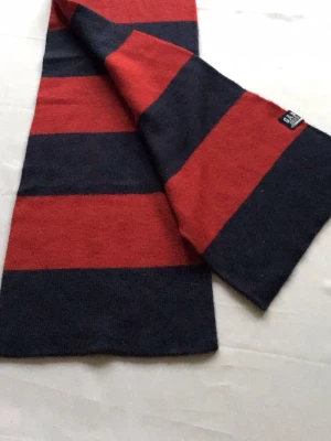 Randig halsduk från GAP i ullmix - Snygg halsduk från GAP med breda röda och mörkblå ränder. Tillverkad i mjuk ullmix som håller dig varm. Klassisk rektangulär form och enkel design som passar till de flesta jackor. GAP-logga på ena kanten för en stilren touch.