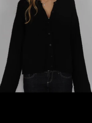 Svart stickad kofta med knappar - En svart stickad kofta med långa ärmar och knäppning framtill. Koftan har en rak passform och är lite croppad, vilket ger en avslappnad vibe. Perfekt att slänga över en t-shirt eller linne för en enkel men snygg look.