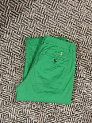 Gröna chinos från Polo Ralph Lauren - Snygga gröna chinos från Polo Ralph Lauren med klassisk passform. Byxorna har en bakficka med knapp och den ikoniska broderade loggan i orange ovanför fickan. Tillverkade i mjuk bomull som känns skön mot huden. Perfekta för dig som vill sticka ut med färg.