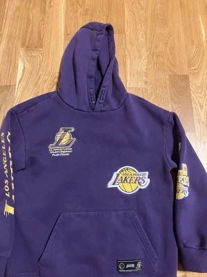 Lila Los Angeles Lakers hoodie - Snygg lila hoodie med Los Angeles Lakers tryck och broderade loggor i gult och vitt. Hoodien har stor huva, magficka och tryck på både bröst och ärmar. Perfekt för dig som älskar basket och vill ha en sportig streetwear-look.