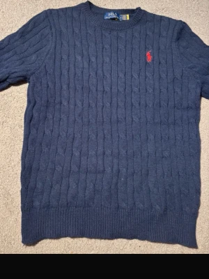 Mörkblå kabelstickad tröja Polo Ralph Lauren - Mörkblå kabelstickad tröja från Polo Ralph Lauren med rund halsringning och röd broderad logga på bröstet. Tröjan har ribbade muddar vid ärmslut och nederkant. Klassisk och tidlös design i mjukt material.