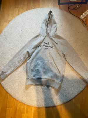 Grå hoodie från Peak Performance - Säljer en grå hoodie från Peak Performance med svart logga på bröstet. Klassisk känguruficka framtill, ribbade muddar och justerbar huva med snören. Mjuk insida i bomullsblandning, perfekt för chill dagar.