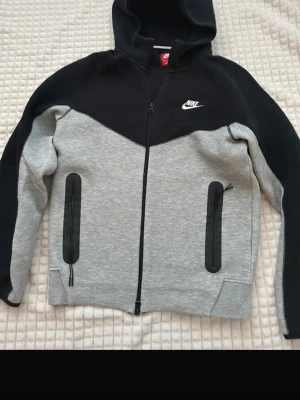 Nike tech  - Snygg hoodie från Nike i svart och grått med dragkedja framtill och två fickor med svarta dragkedjor. Klassisk Nike-logga på bröstet och huva. Perfekt för en sportig och avslappnad stil.