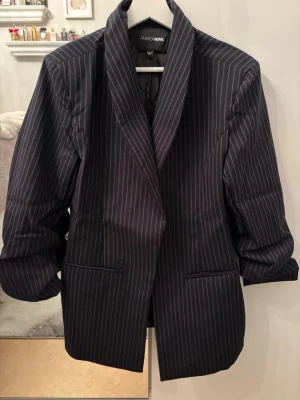 Mörkblå randig kavaj Fashion Nova - Snygg mörkblå kavaj från Fashion Nova med vita tunna kritstrecksränder. Klassisk passform med breda slag och två framfickor. Perfekt för en stilren och trendig look. Materialet känns slätt och har en lätt glans.