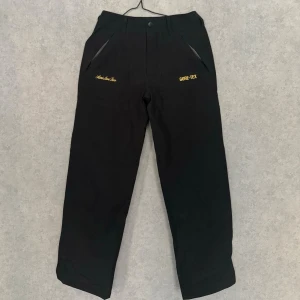 Aimé Leon Dore/Goretex Trousers - Svarta byxor från Aimé Leon Dore med gul brodyr och GORE-TEX-detalj. Byxorna har raka ben, dragkedjeförsedda fickor och bälteshällor. Materialet är slitstarkt och vattenavvisande, perfekt för en streetwear-look eller skidbacken