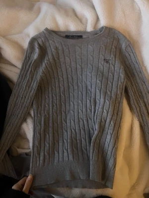 Grå ribbad tröja från GANT - Säljer en grå kabelstickad tröja från GANT i mjuk bomull. Tröjan har rund halsringning, långa ärmar och diskret GANT-logga broderad på bröstet. Klassisk och enkel design som passar till det mesta. Näshan aldrig använd, i XS