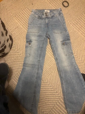 Ljusblå cargobyxor i denim - Säljer ett par ljusblå cargobyxor från Cubus i klassisk denim. Byxorna har flera praktiska fickor på sidorna och en relaxed passform med lätt utsvängda ben. Perfekta för en chill och trendig look.