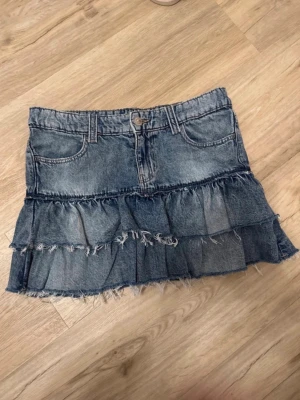 Jeanskjol med volanger och råa kanter - Snygg kort jeanskjol med volangdetaljer och råa, fransiga kanter. Kjolen har klassisk femficksdesign och bälteshällor. Perfekt för dig som gillar en trendig och avslappnad stil.