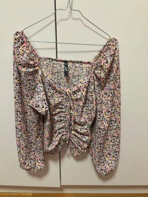 Blommig omlottopp stl 30 - Söt blommig omlottopp med volangdetaljer och snörning framtill. Toppen har långa ärmar och offshoulder-design. Färgerna går i ljusrosa, gul, svart och vit, perfekt för vår och sommar.