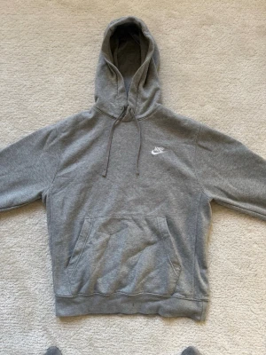 Grå hoodie från Nike med huva - Klassisk grå hoodie från Nike med vit logga på bröstet. Hoodien har stor känguruficka framtill, justerbar dragsko i huvan och mjukt, borstat innerfoder. Perfekt för en avslappnad och sportig stil.