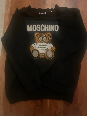 Svart Moschino hoodie med nalle - Säljer en svart hoodie från Moschino med ett stort broderat motiv av en nallebjörn på bröstet och texten 'This is not a Moschino toy'. Hoodien har en avslappnad passform och är tillverkad i mjukt material. Perfekt för dig som gillar statement-plagg och streetwear.