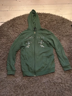 Grön bling lucid rhinestone zip hoodie - Grön hoodie med dragkedja och huva. På bröstet finns två ängeltryck och texten 'BLING LUCID' med svarta och gråa rhinestone paljetter. Hoodien har långa ärmar och två fickor framtill. Perfekt för en chill och avslappnad stil. Läs deras hemsida om hur man ska tvätta den. Passar mer som S!👍🤗
