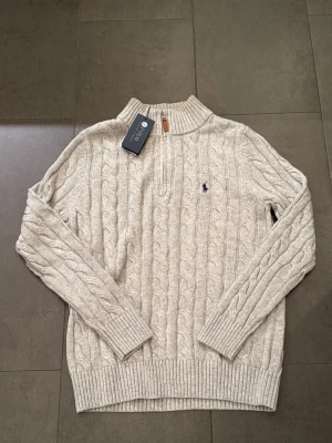 Beige kabelstickad tröja Polo Ralph Lauren - Kabelstickad beige tröja från Polo Ralph Lauren med halv dragkedja och ribbade muddar. Tröjan har lång ärm och klassisk broderad logga på bröstet. Perfekt för lager-på-lager och snygg till jeans eller chinos.