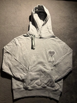 Grå hoodie med broderad logga - Snygg grå hoodie från AMI Paris med stor huva, dragsko och känguruficka. På bröstet sitter en broderad logga i form av ett hjärta med bokstaven A. Tillverkad i mjuk bomullsblandning som känns skön mot huden. Perfekt för en chill och avslappnad stil.