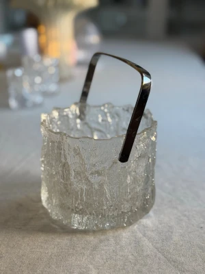 Isbehållare i glas med metallhandtag littala - Säljer en stilren isbehållare i klart glas med frostad, isliknande struktur och vågig kant. Behållaren har ett avtagbart handtag i blank metall som ger en modern touch. Perfekt till drinkbordet eller som dekoration. Littala 