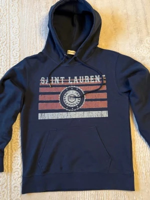 Saint Laurent Hoodie  - Säljer en snygg Saint Laurent hoodie i mörkblå färg med tryck framtill. En riktigt clean och stilren hoodie som passar till allt.  ✅ Bekväm passform ✅ Tjock och bra kvalitet ✅ Klassisk design med tryck ✅ Perfekt för vardag  Använd men fortfarande i bra skick (se bilder för detaljer). Inga större defekter.  📦 Skickas snabbt 💬 Hör av dig vid frågor eller fler bilder!